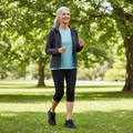 An older woman with gray hair, dressed in a blue shirt, black leggings, and a dark jacket, smiles while enjoying a sunny walk in the park wearing Skechers Arch Fit 2.0 Slip In WIDE FIT Lace Up Easy Chic Black shoes., 50572, 198376162054, 198376162078, 198376162092, 198376162115, 50581