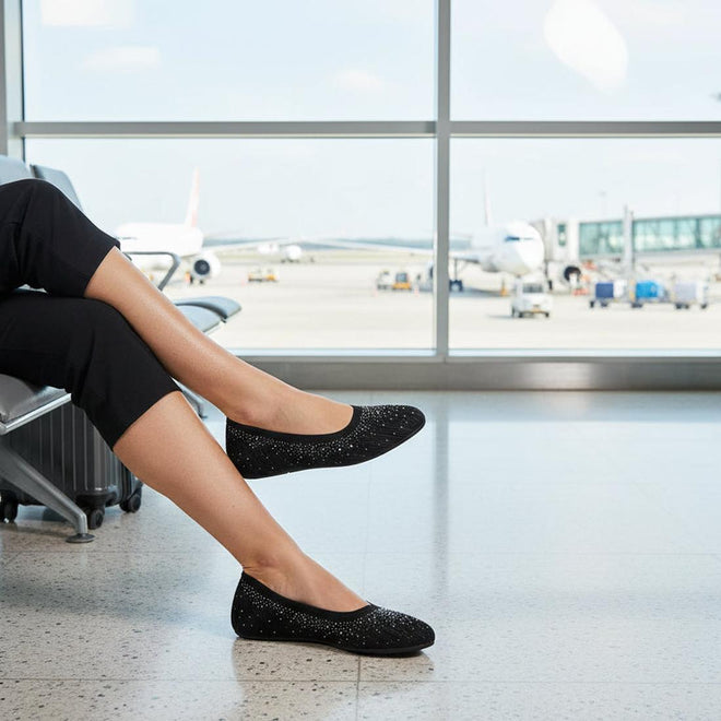 A person sits in an airport terminal with legs crossed, wearing black Womens Skechers Cleo 2.0 Glitzy Daze Ballet Flats and black pants, with airplanes and activity visible through the large windows in the background., 50212, 196989721118, 196989721132, 196989721156, 196989721170, 50221