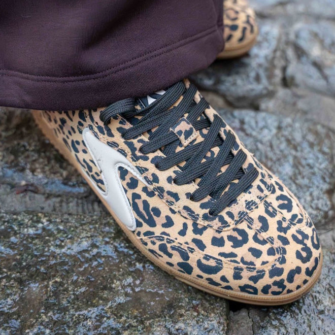 A close-up of someone wearing the Skechers Womens HotShot On The Prowl Leopard Suede Sneaker with brown and black leopard print, Air-Cooled Memory Foam, black laces, and white side detail, standing on stone pavement., 52252, 199025014359, 199025014373, 199025014380, 199025014397, 199025014410, 52257
