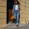 A woman in a gray blazer, white top, and wide-leg jeans walks confidently outside by a yellow wall. She carries a small orange bag, wears sunglasses, and completes her look with Woden Line Women's 3 Strap Sandal Dark Olive., 5713326432305, 5713326432312, 5713326432329, 5713326432336, 5713326432343, 5713326432350, 5713326432367