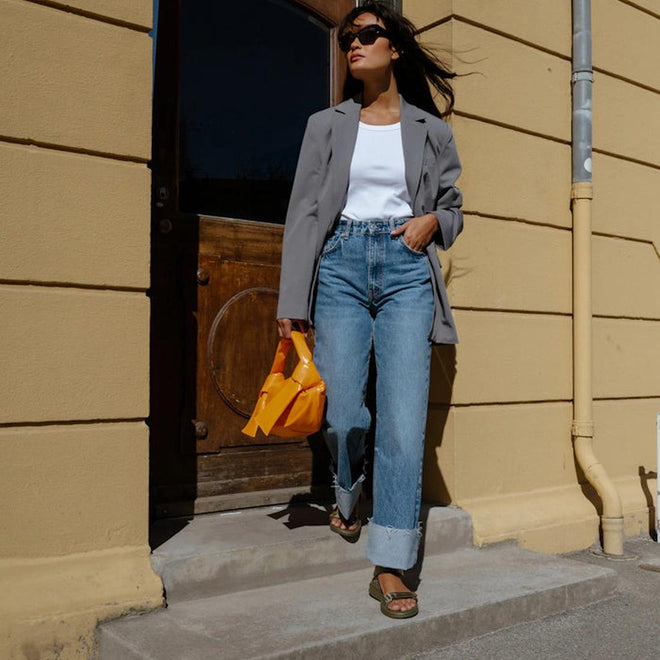 A woman in a gray blazer, white top, and wide-leg jeans walks confidently outside by a yellow wall. She carries a small orange bag, wears sunglasses, and completes her look with Woden Line Women's 3 Strap Sandal Dark Olive., 5713326432305, 5713326432312, 5713326432329, 5713326432336, 5713326432343, 5713326432350, 5713326432367