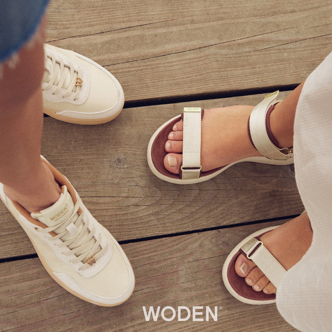 Two people stand on a wooden deck, one wearing ivory Woden Line Sneak Ivory Lace Up Sneaker shoes and the other wearing beige strappy sandals.