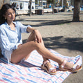A woman sits on a striped blanket wearing comfortable pink and white Woden Line Women’s 3 Strap Sandal Blush Multi sandals with thick, adjustable three-strap soles.