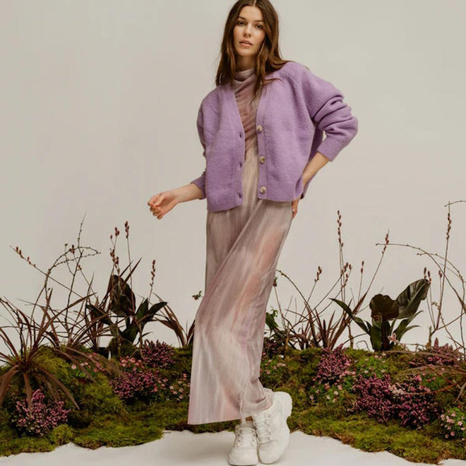 A woman stands on moss and plants in a pastel dress, lavender cardigan, and Woden SIF Reflective Women's Sneaker Blanc de Blanc White. The plain, light background adds to the soft, natural vibe., 36107, 5713326428612, 5713326428629, 5713326428636, 36110