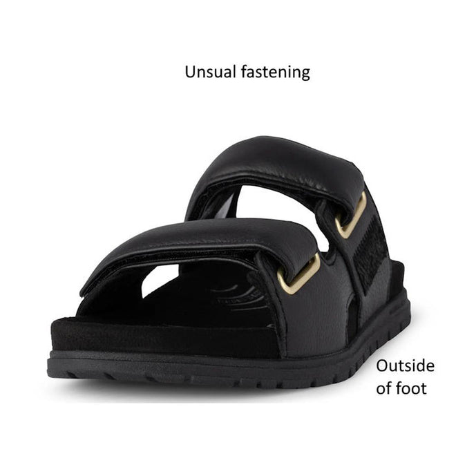 The Woden Lisa Women's Leather 2 Strap Sandal in black is shown from the front, featuring a unique fastening design, chunky buckle straps, and gold hardware. Text labels point out the distinctive fastening and outside of the foot., 5713326518276, 5713326518283, 5713326518290, 5713326518306, 5713326518313, 3935713326518320, 5713326518269