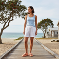 A woman stands on a wooden boardwalk by the beach, wearing a blue striped tank top, white shorts, and Crocs Womens Getaway PLATFORM Flip Sandal in Sunstone Orange. Trees and beach cabins surround her, with the ocean and sky behind., 49910, 49911, 198445274329, 198445274336, 198445274343, 49915, 49916