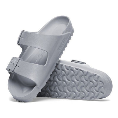 A pair of Birkenstock Arizona EVA Stone Coin Grey Regular Width sandals with two adjustable straps, textured soles, and an anatomically shaped footbed. One stands upright, the other lies flat, highlighting their waterproof design.