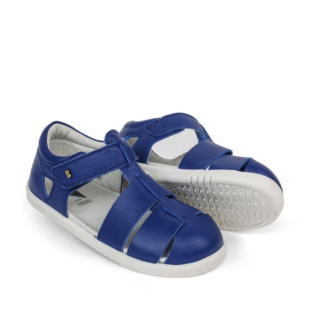 The Bobux Iwalk IW Quick Dry Tidal Sandal 634408 Blueberry is a pair of unisex kids’ blue quick-dry leather sandals with closed toes, velcro straps, and white rubber soles. One sandal stands upright; the other lies sideways, showing its textured sole.