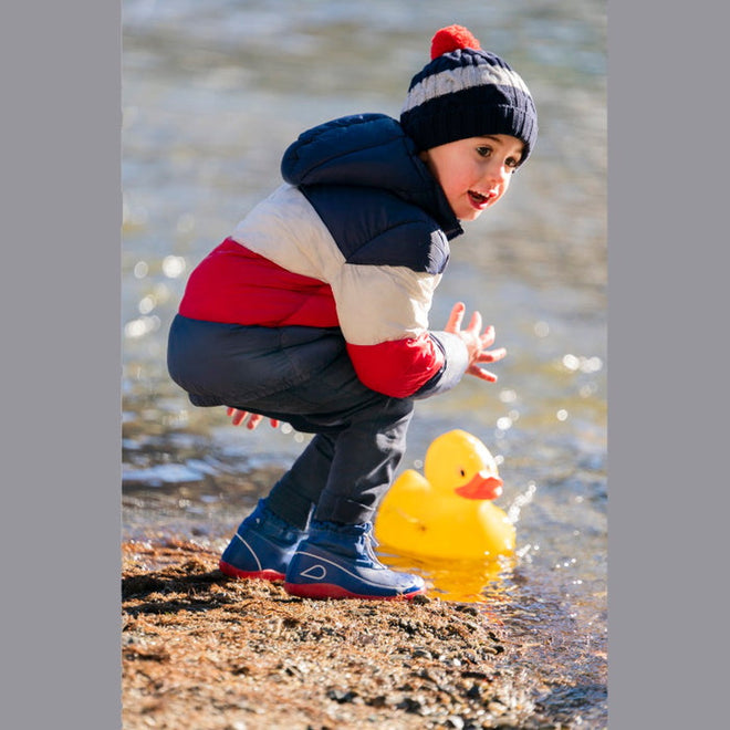 A young child in a striped winter jacket, hat, and blue boots squats by the water's edge, playing with a large yellow rubber duck—perfect for showcasing the Bobux Kid+ Splash Boot Waterproof Blueberry 839003., 9420068527911, 9420068527928, 9420068527935, 9420068527942, 9420068527959, 9420068527966, 9420068527973