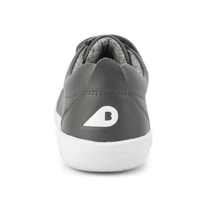 A close-up rear view shows the Bobux Kids + Plus Grass Court Smoke Grey Velcro Trainers 832446, featuring gray leather, a white sole, and a white B logo on the heel—perfect street shoes for kids—set against a plain white background., 9420068518025, 9420068518032, 9420068518049, 9420068518056, 9420068518063, 9420068518070, 9420068518087