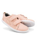 The Bobux KP Kid Plus Grass Court Seashell Pink 832448 are light pink kids trainers featuring a leather upper, white soles, double Velcro straps, heart-shaped perforations, and a textured sole. One shoe stands upright while the other lies on its side.