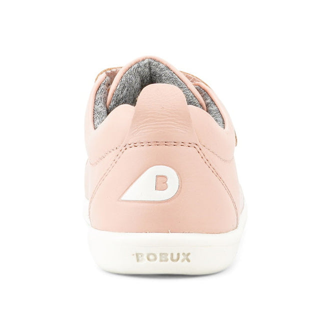 A close-up rear view of the Bobux KP Kid Plus Grass Court Seashell Pink 832448 kids trainer shows a pale pink leather upper, white sole, gray lining, double velcro straps, a white B logo on the heel, and BOBUX text on the back., 9420075792579, 9420075793040, 9420075793125, 9420075794306, 9420075791602, 9420068517837, 9420068517844