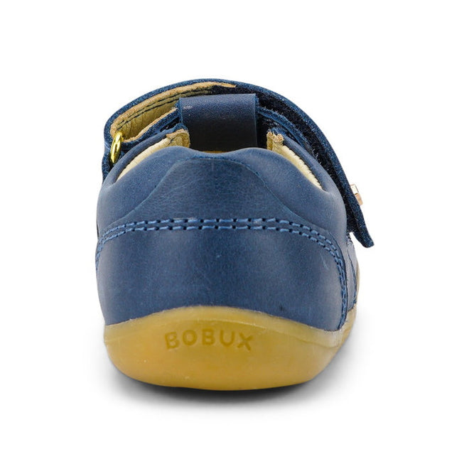 A close-up rear view of the Bobux Step Up T-Bar Louise Midnight Blue 728330 shows its small blue leather upper, yellow stitching, tan rubber sole, and BOBUX embossed on the heel., 9420068521810, 9420068521827, 9420068521834