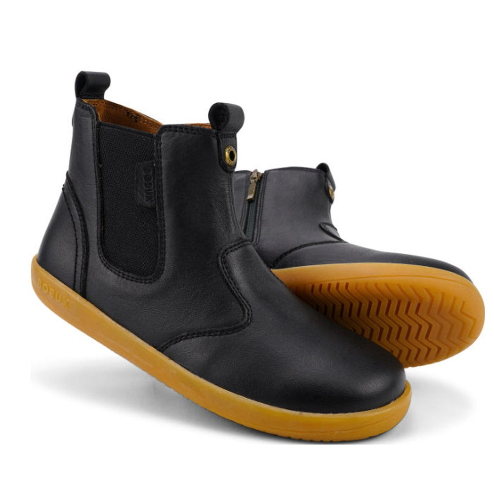 The Bobux Kid+ Jodhpur Boot Black 830016Z is a pair of unisex leather ankle boots featuring side elastic panels, pull tabs, and light brown rubber soles. One boot stands upright while the other displays the tread pattern.