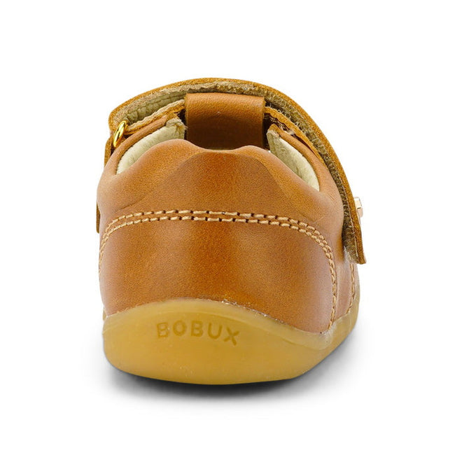 A rear view of the Bobux Step Up T-Bar Louise Tan Caramel 728318 shows its small brown leather design, soft rubber sole with BOBUX embossed on the heel, white stitching, and an adjustable velcro strap., 9420062092132, 9420062092149, 9420062092156, 9420062092163