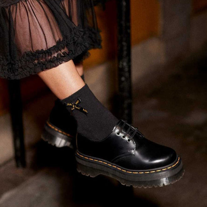 A person sits cross-legged wearing a sheer black skirt, Dr Martens Ankle Bow Cotton Blend Socks in black, and shiny black Airwair platform Oxford shoes with yellow stitching., 190665563641, 190665563634