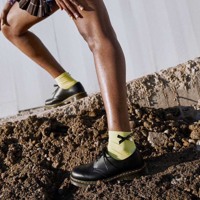 Someone wearing Dr Martens Ankle Bow Cotton Blend Socks in Lemon with black shoes and a plaid skirt steps onto rocky ground outdoors., 190665558562, 190665558555