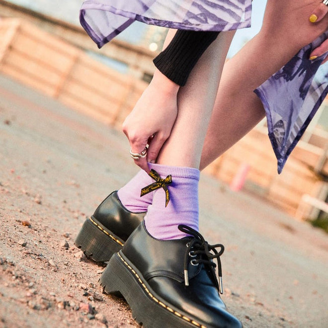 Someone adjusts Dr Martens Ankle Bow Cotton Blend Socks in lilac (Small/Medium up to UK7, Medium/Large above UK7) with a yellow and black bow, styled with black platform shoes and a sheer purple skirt on sandy ground., 190665558609, 190665558593