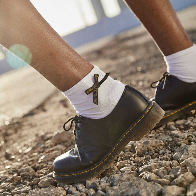 Close-up of a person wearing Dr Martens Ankle Bow Cotton Blend Socks in white, paired with black leather shoes with yellow stitching. These machine washable socks are available in Small/Medium (up to UK7) and Medium/Large (above UK7)., 190665563665, 190665563658
