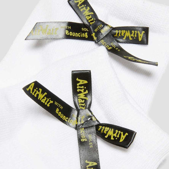 A close-up of Dr Martens Ankle Bow Cotton Blend Socks in white, featuring black ribbon bows with yellow “AirWair with Bouncing Soles” text—a cute, machine washable ankle style available in Small/Medium up to UK7 and Medium/Large above UK7., 190665563665, 190665563658