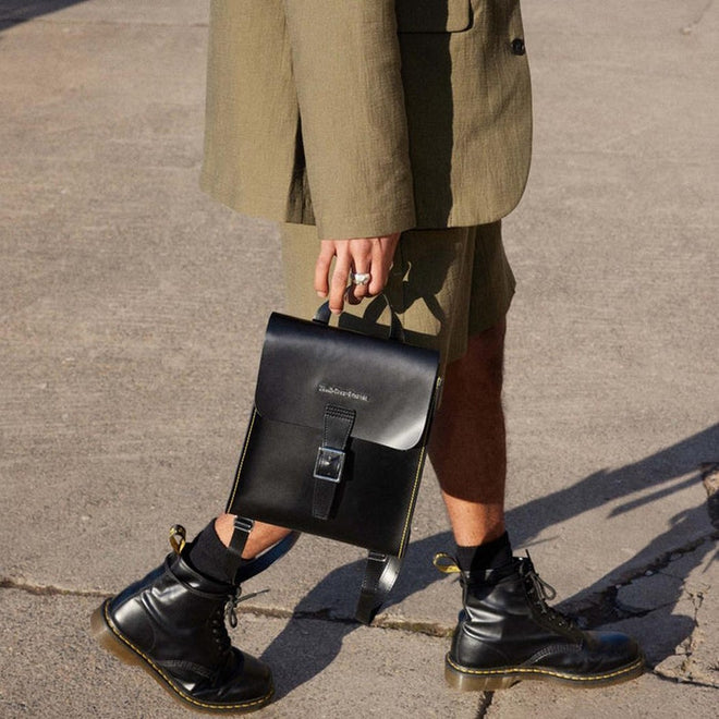 190665192032  
Side view of a person walking carrying the dr martens backpack in her hand by the top strap, showing the size relative to a person.