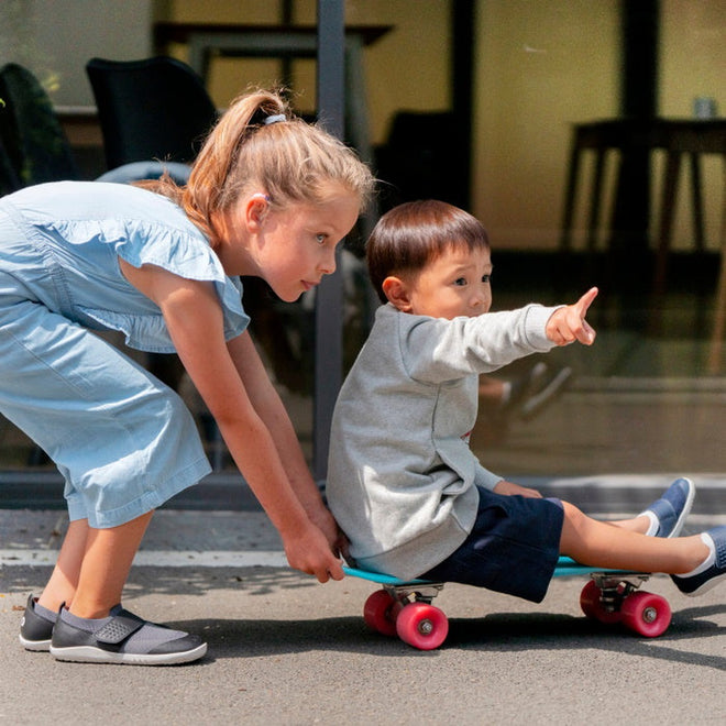 A young girl in a light blue outfit, wearing Bobux Iwalk Lo Dimension III Blue Navy Trainer 638506 Unisex shoes, pushes a boy on a skateboard with pink wheels. The boy, in a gray sweater and shorts, points ahead as they play outdoors near glass., 9420062025925, 9420071311699, 9420068531536, 9420071311712, 9420068531550