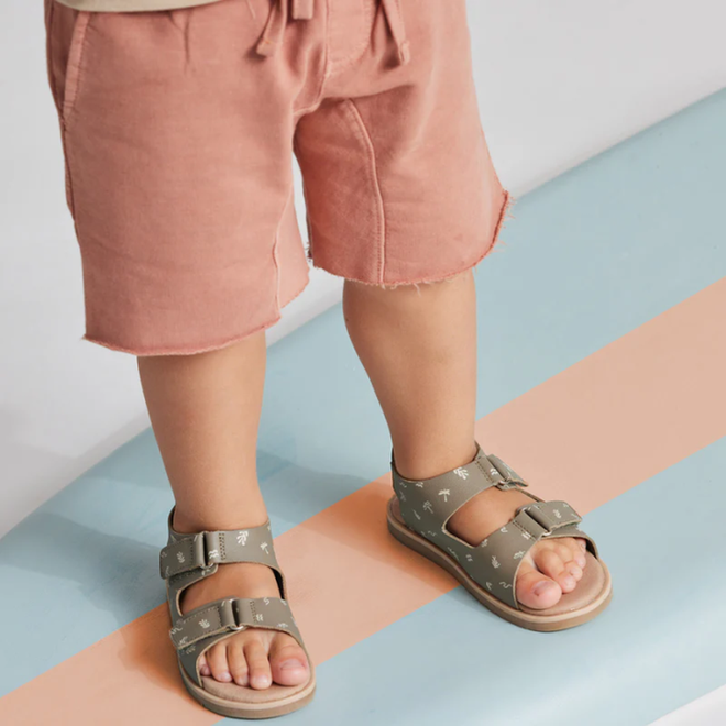 A young child in light pink shorts and Pretty Brave Wilder Reef Green Kids Sandals (sizes 29-31, not returnable), featuring white patterns and adjustable velcro straps, stands on a pastel blue and peach striped surface., 9421036359503, 9421036359510, 9421036359527, 25284, 25285, 25286, 25287