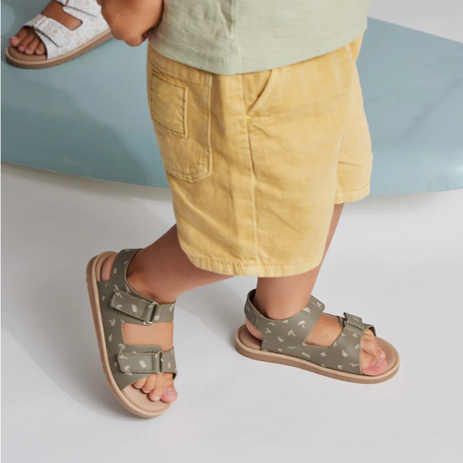 A young child in yellow shorts wears Pretty Brave Wilder Reef Green Kids Sandals (29-31, NOT RETURNABLE) with adjustable velcro straps on a light blue surface. Another child's feet in white kids sandals are visible in the background., 9421036359503, 9421036359510, 9421036359527, 25284, 25285, 25286, 25287