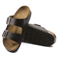 a pair of Birkenstock Arizona Smooth Leather Dark Brown Regular Width with double straps, one over the foot and one over the toes with buckle adjustment. Staps are linked at the sides of the foot. Leather thick straps and a suede leather footbed. Also showing the out sole of the left foot lying down.
