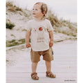 A young child stands on sandy ground, wearing a cream boho tee, tan shorts, and Bobux Step Up Todders Sandal Roam 729204B in Caramel. The child smiles and looks upward with greenery and sand in the background., 15014, 9420062081464, 9420062081471, 9420062081488