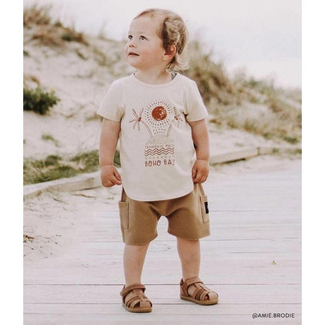 A young child stands on sandy ground, wearing a cream boho tee, tan shorts, and Bobux Step Up Todders Sandal Roam 729204B in Caramel. The child smiles and looks upward with greenery and sand in the background., 15014, 9420062081464, 9420062081471, 9420062081488
