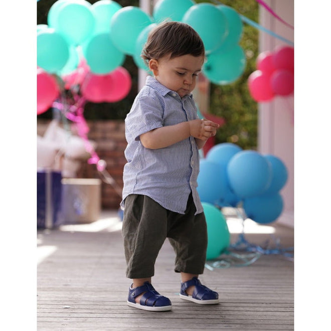 A young boy in a blue striped shirt, dark pants, and Bobux Step Up US Tidal Navy Closed Toe Sandal 732503 stands on a wooden floor with colorful balloons and party decorations in the background., 9420068502574, 9420068502581, 9420068502598, 9420068502604