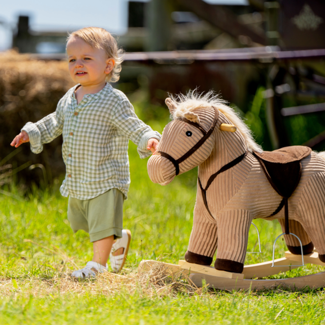 A young child walks outdoors on grass, wearing a light checked shirt, shorts, and Bobux iwalk Cross Jump Sandal White Closed Toe 636706 with Velcro fastening. The child holds the mane of a plush rocking horse under bright sunlight., 9420062083949, 9420062083956, 9420062083963, 9420062083970, 9420062083987