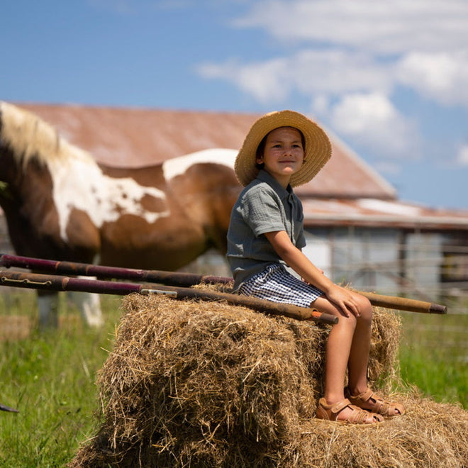 A young child in a straw hat and Bobux Kid+ Plus Roam Caramel Closed Toe Sandal (830506A) sits on a hay bale on a sunny farm, with a brown and white horse and rustic barn in the background., 9420062049952, 9420062049969, 9420062049976, 9420062049983, 9420062049990, 9420062050002, 9420062050019