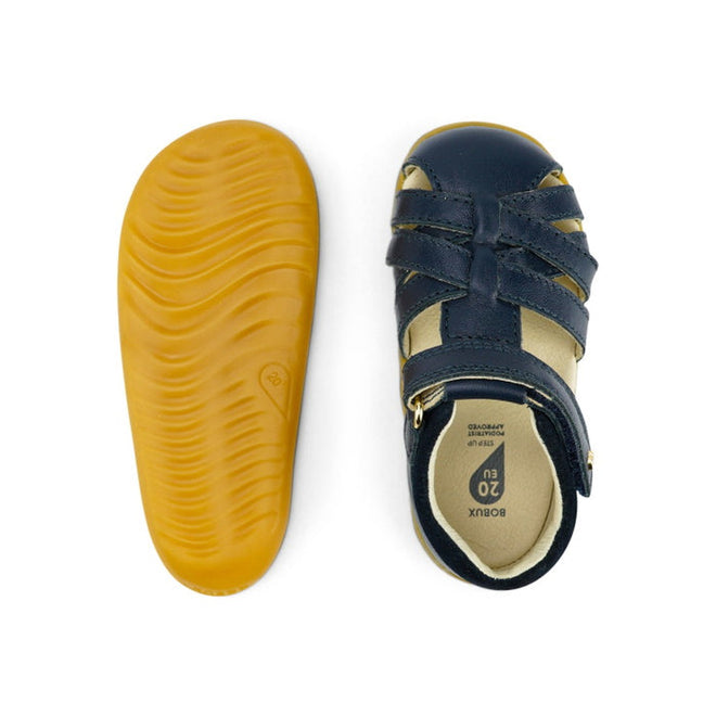 A pair of Bobux Step Ups Cross Jump Navy Closed Toe Sandal unisex; one shoe displays dark blue straps and a beige interior from the top, while the other shows its flexible, textured yellow sole from the bottom., 9420062083567, 9420062083574, 9420062083581, 9420062083598