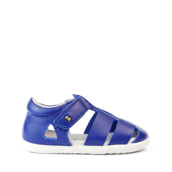 The Bobux Step Up US Tidal Blueberry Closed Toe Sandal 732501 is a single blue toddler sandal with a closed toe and heel, cutout details, velcro strap, and white sole—ideal as first walker shoes—shown on a white background., 9420068502352, 9420068502369, 9420068502383, 9420068502376