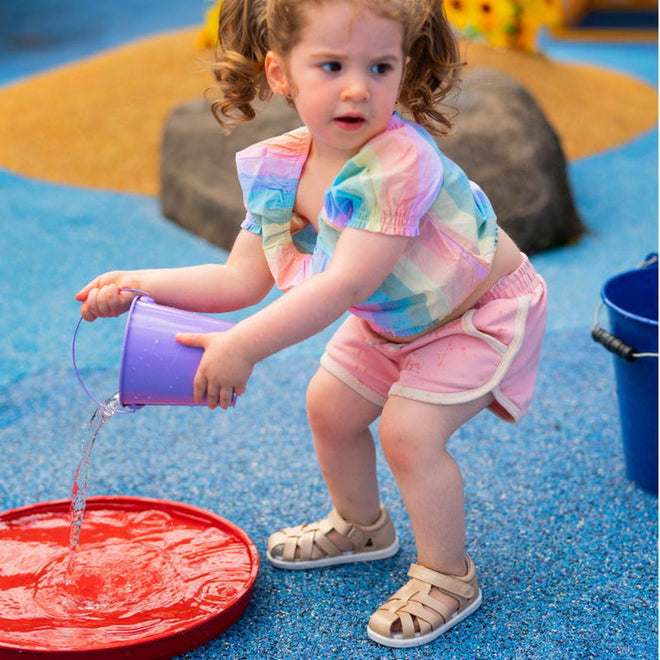 A young girl with curly hair, wearing a colorful shirt, pink shorts, and Bobux Step Up SU Tropicana II Pale Gold 732311 sandals, crouches at a splash pad as she pours water from a purple bucket onto the red surface., 9420068529915, 9420068529922, 9420068529939, 9420068529908