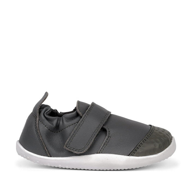 The Bobux Xplorer XP Go Smoke Grey First Walker Crawler 501002B is a dark gray children's shoe with a white flexible sole, velcro strap, and reinforced toe cap, made from premium soft leather and shown in profile against a white background., 9420062079744, 9420062079751, 9420062079768, 9420062079775, 9420062079782