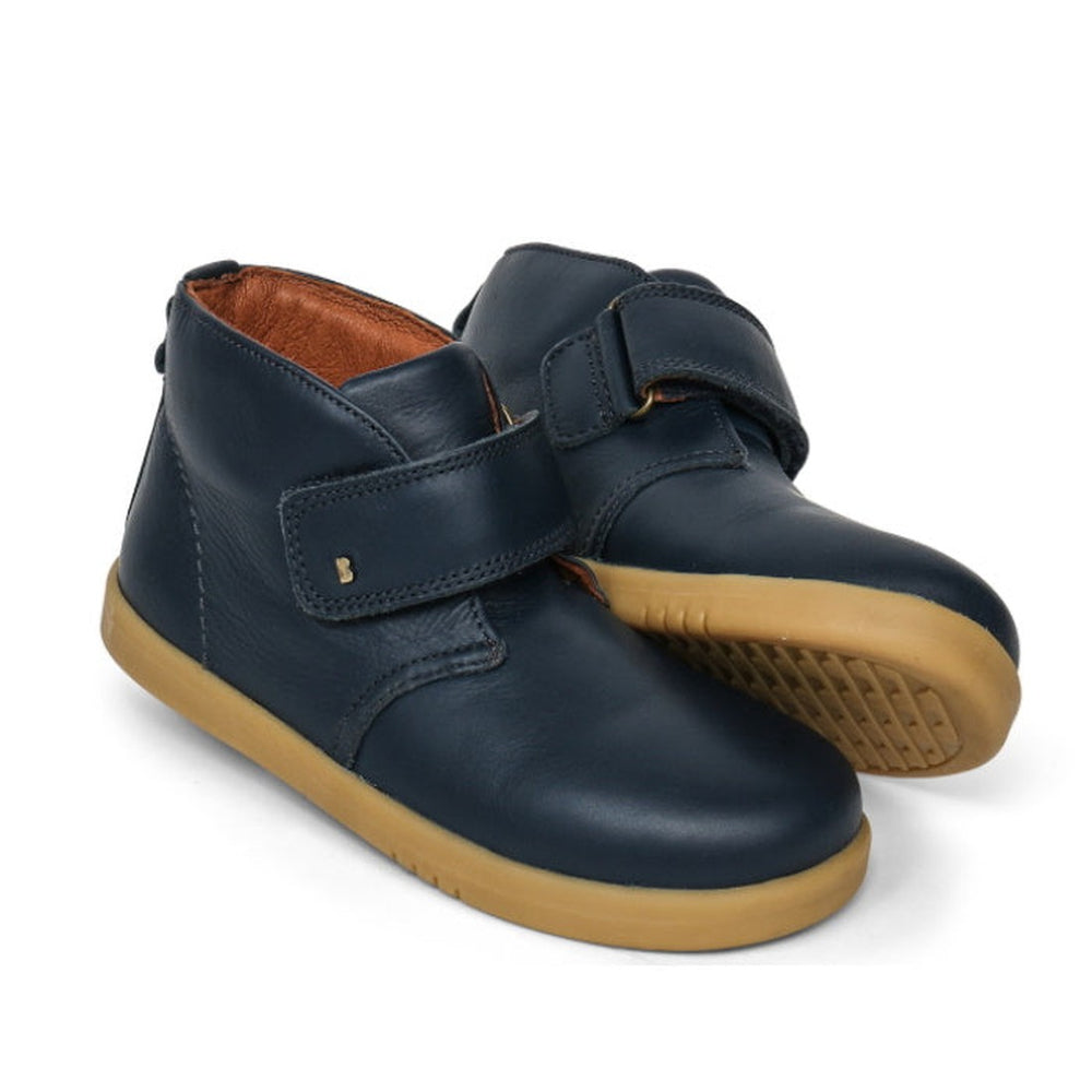 The Bobux Kid+ Desert Boot Navy 830304Z is a unisex children's high-top navy blue leather boot with tan rubber soles and a wide Velcro opening, shown on a white background.