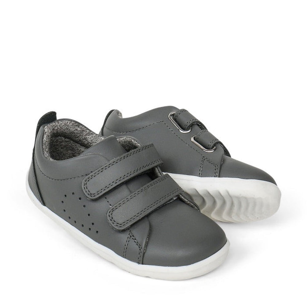 A pair of Bobux Step Up First Shoes Grass Court Trainer 728913 in Smoke Grey, featuring soft leather, white soles, and Velcro straps. One shoe is slightly behind and angled to display the side and front—ideal for first walkers.