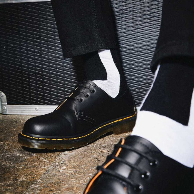 A person in black leather shoes with yellow stitching, black pants, and Dr Martens Socks Double Doc Sock Unisex Black White for blister protection stands on a textured concrete floor beside a black ridged case., 190665377309, 190665377293