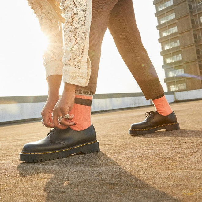 A person wearing Dr Martens Organic Socks Athletic Logo Beige and black lace-up shoes bends to adjust their pants on a rooftop, sunlight shining behind them and a tall building in the background., 190665558487, 190665558470