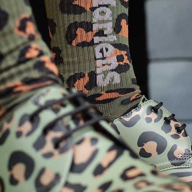 Close-up of a person wearing Dr Martens Socks Vertical Logo Unisex Camo Khaki—bold cotton-blend socks in green camo with a vertical logo, paired with matching lace-up shoes featuring a similar pattern., 190665515374, 190665515367