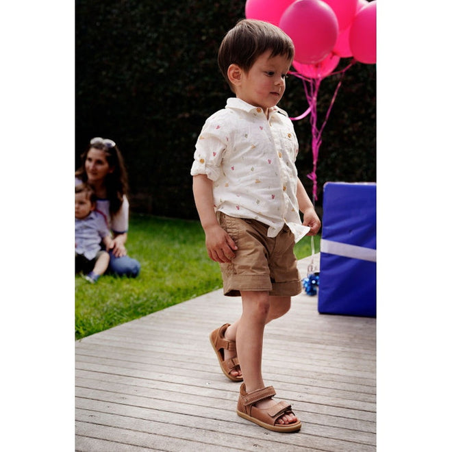 A young boy wearing a white shirt, tan shorts, and Bobux IWalk Driftwood Sandal Caramel Open Toe 633606A walks on a wooden deck. Pink balloons, a large blue gift box, and two people on the grass are in the background., 9420062048085, 9420062048078, 9420062048092, 9420062048108, 9420062048115