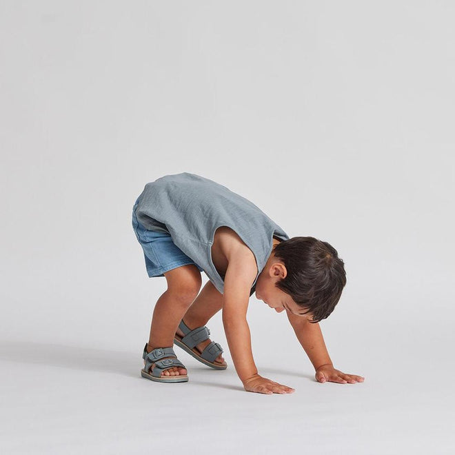 A young child in a gray sleeveless shirt, denim shorts, and Pretty Brave Alex Leather Sandal Seaform Blue (sizes 29-33, CLEARANCE NOT RETURNABLE) bends over with hands on the ground against a plain light background., 9421036357684, 9421036357691, 9421036357707, 9421036357714, 9421036357721